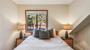 Ein Schlafzimmer mit einem großen Bett, zwei Nachttischen, zwei Lampen und einem Fenster mit Blick auf Bäume und ein Gebäude.
