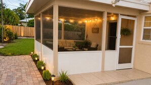 A covered patio with string lights, a seating area with cushions, and a small garden with plants and a pathway. - Mercedes Isle Retreat | 3BR + Private Fenced Yard (Stuart)