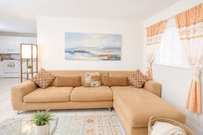 A beige sofa with patterned cushions, a painting of a landscape, and a window with curtains.