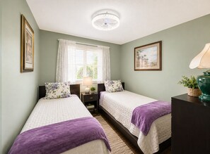 A bedroom with two beds, a nightstand, a lamp, a ceiling fan, and a window with curtains.