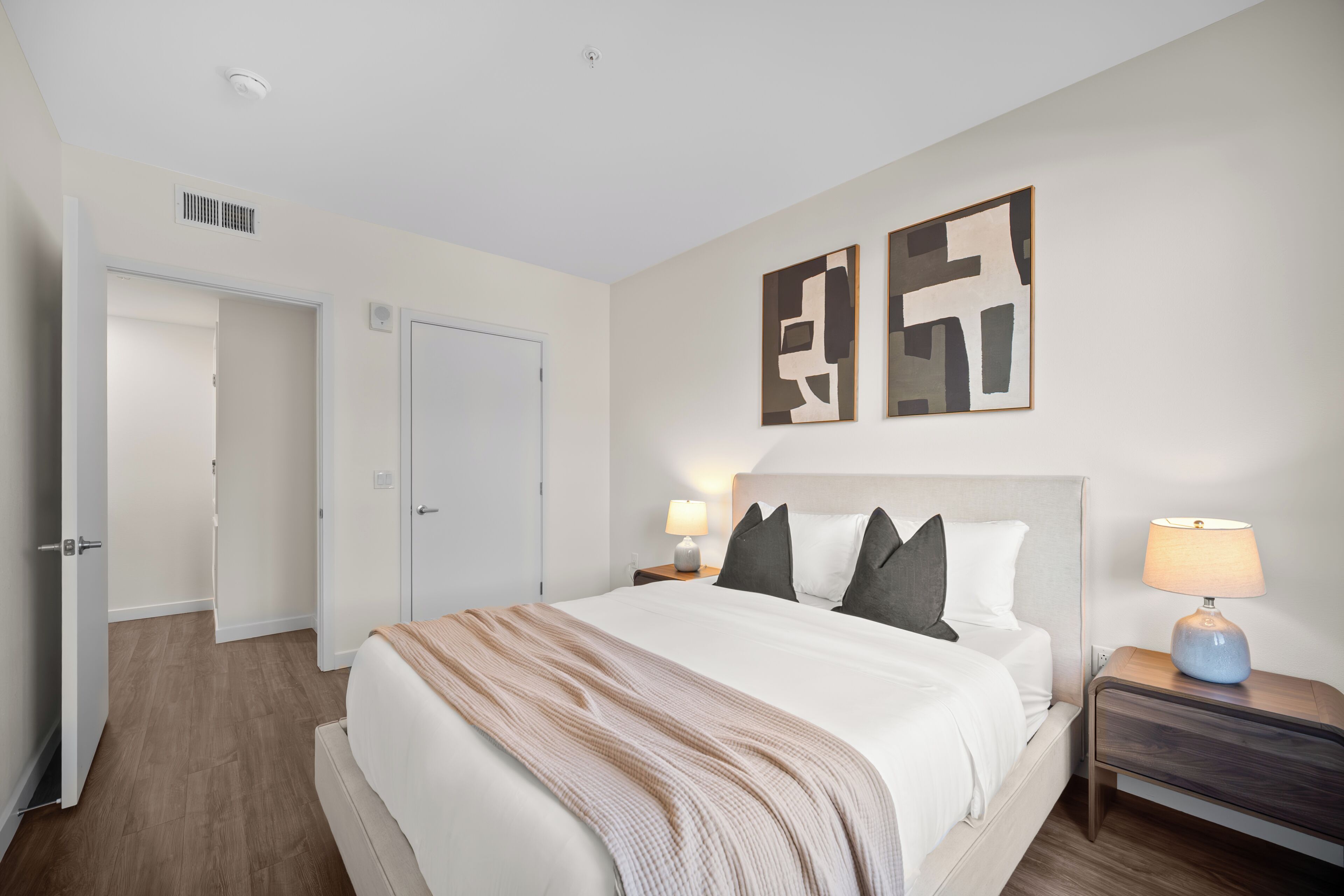 Una habitación de hotel con una cama grande, dos mesitas de noche con lámparas, una puerta y arte abstracto en la pared.
