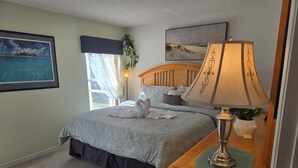A neatly made bed with a grey bedspread and two white towels, a wooden nightstand with a lamp, a framed picture on the wall, and a window with curtains. - Low Rates! Superb 4-bed Pool/Game Room Home Near Disney World Free Wi-fi & More (Kissimmee)