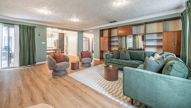Ein großzügiger Wohnraum mit einem grünen Sofa, einem grauen Sessel und einem Holztisch. Es gibt einen Holzschrank mit Regalen und ein großes Fenster mit Vorhängen.