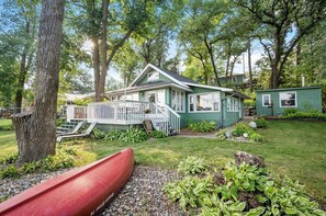 Une maison verte avec un porche, entourée d’arbres et à côté d’un canoë rouge.