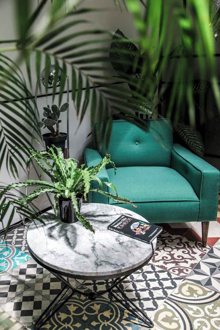 A teal armchair, a marble table, and potted plants in a room with patterned floor tiles.