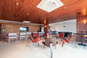 A modern interior with a wooden ceiling, a TV mounted on the wall, and a seating area with red chairs and a round table. - Beachfront Building With Pool and gym - Arus (Salvador)