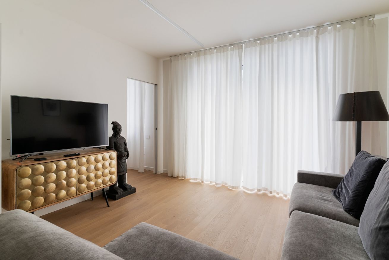 Una sala de estar con televisor de pantalla plana, un mueble de madera de diseño esférico, una estatua, un sofá gris y un ventanal con cortinas translúcidas.