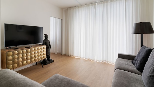Un salon doté d’un téléviseur à écran plat, d’un meuble en bois au design sphérique, d’une statue, d’un canapé gris et d’une grande fenêtre ornée de rideaux légers.