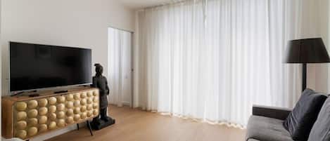 Una sala de estar con televisor de pantalla plana, un mueble de madera de diseño esférico, una estatua, un sofá gris y un ventanal con cortinas translúcidas.