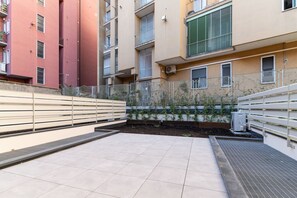 Un patio con piso de baldosas, cercado por una valla metálica, y un edificio con varios balcones y ventanas.