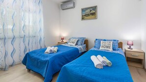 Ein Hotelzimmer mit zwei Betten, jeweils mit blauer Bettdecke und Kissen, einem kleinen Nachttisch mit Lampe, einer Wandklimaanlage und einem gerahmten Bild über den Betten.