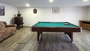Ein Billardtisch in einem Raum mit Holzboden, einer Couch, einer Dartscheibe und einem gerahmten Bild an der Wand.
