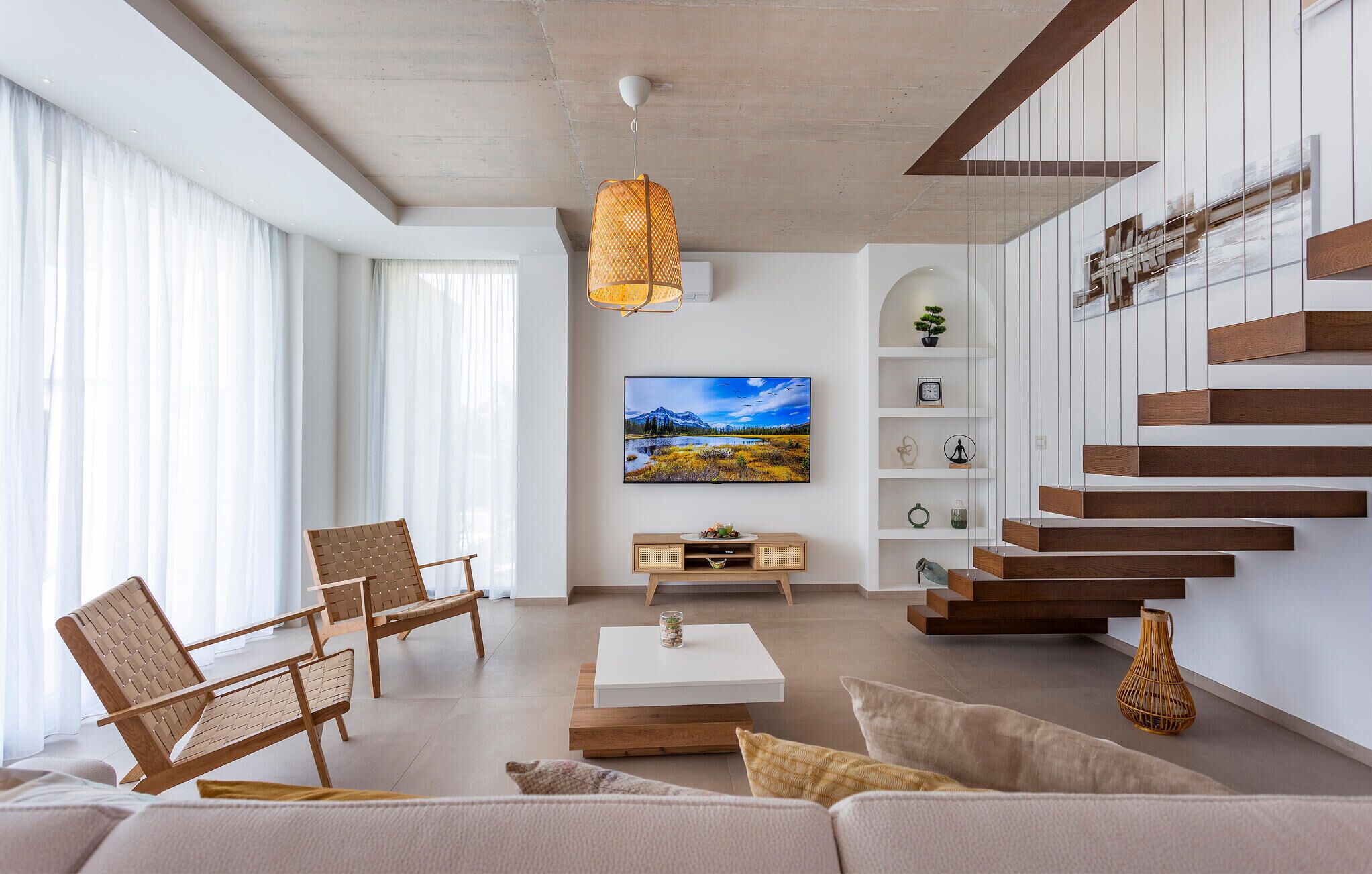 Una sala de estar moderna con un televisor de pantalla plana grande, muebles de madera, una escalera y vistas a un paisaje montañoso.