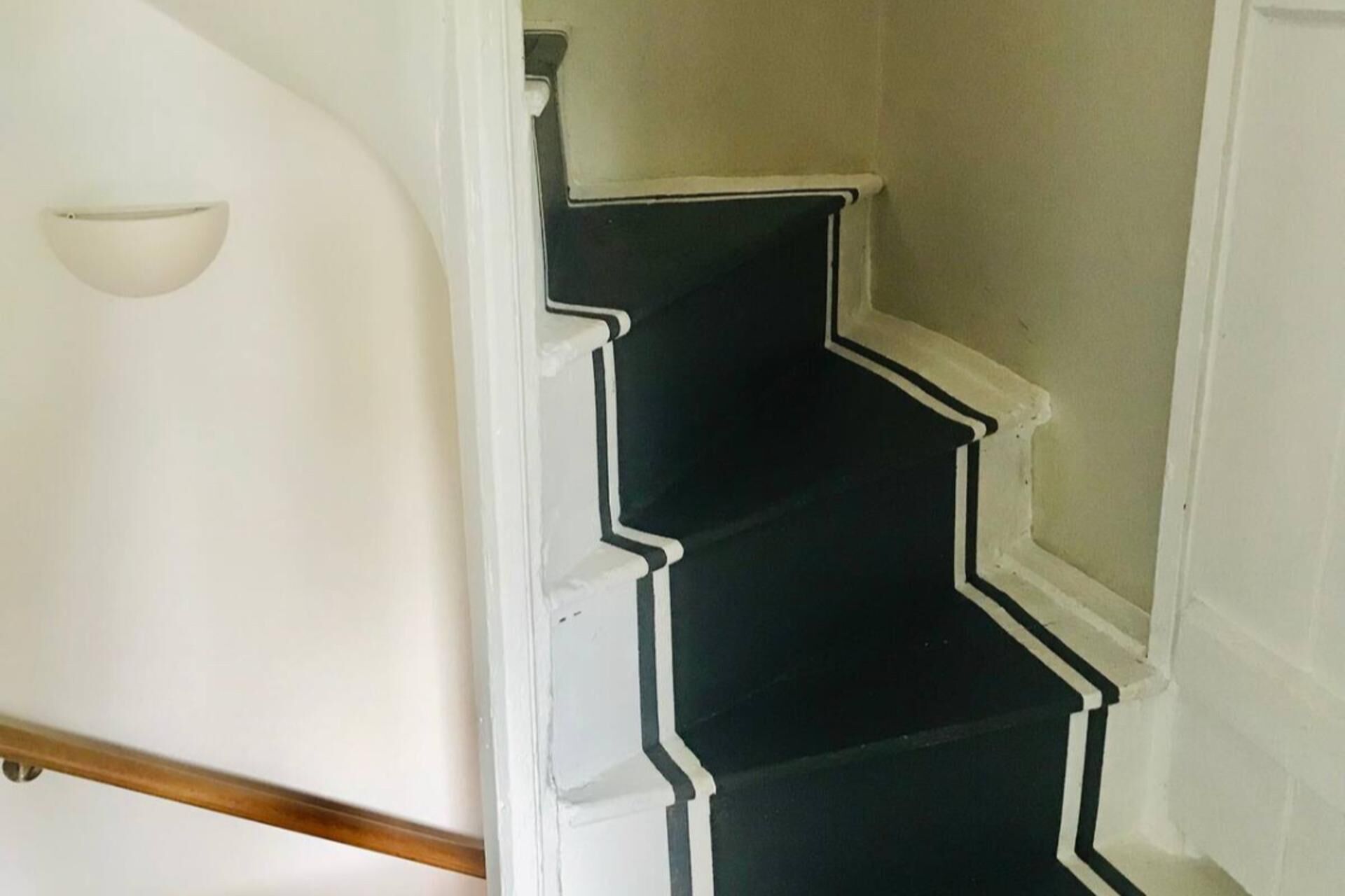 A staircase with black steps and white edges, leading to a landing with a white wall and a light fixture.