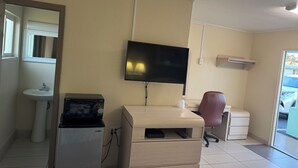 A room with a flat-screen TV mounted on the wall, a wooden desk, a red office chair, a white sink, and a compact refrigerator. - American Inn (Cocoa)
