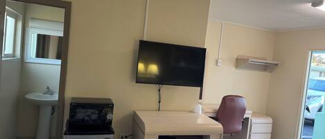 Een kamer met een wandgemonteerde flatscreen-tv, een houten bureau, een rode bureaustoel, een witte wastafel en een compacte koelkast.