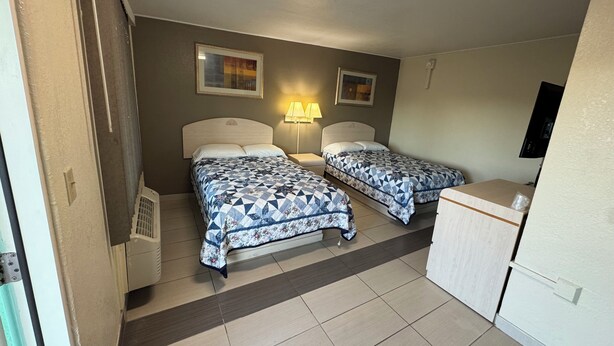 A room with two beds, a desk, and a chair. There are two wall-mounted lamps and a ceiling light. - American Inn (Cocoa)