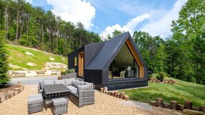Una cabaña moderna con techo inclinado, un ventanal grande y mobiliario exterior.