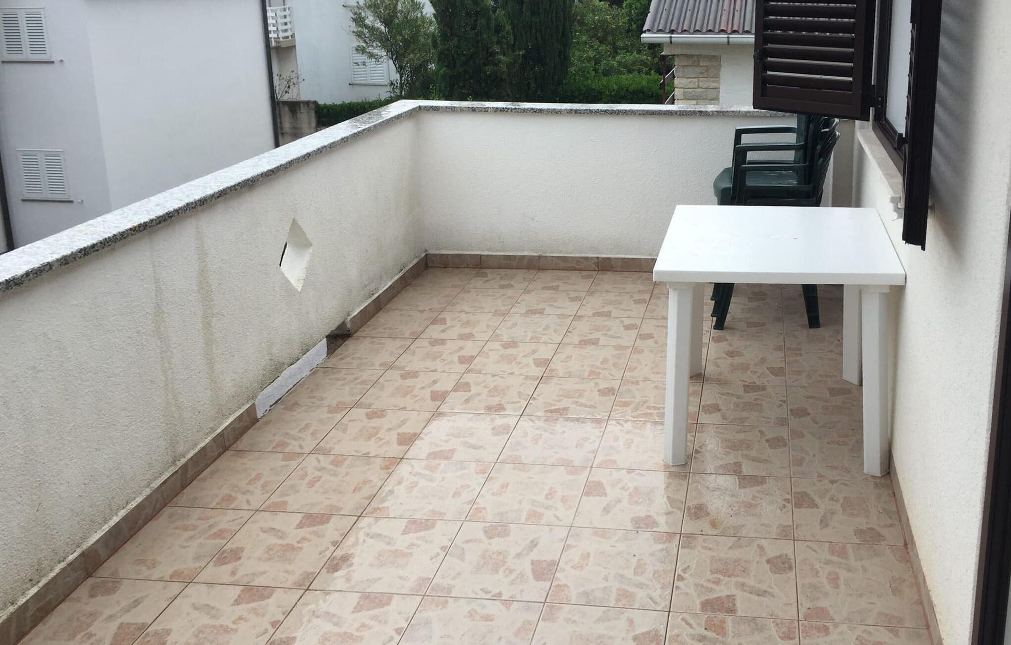 Een klein balkon met een witte tafel en stoelen, een betegelde vloer en een witte muur met een raam en luiken.