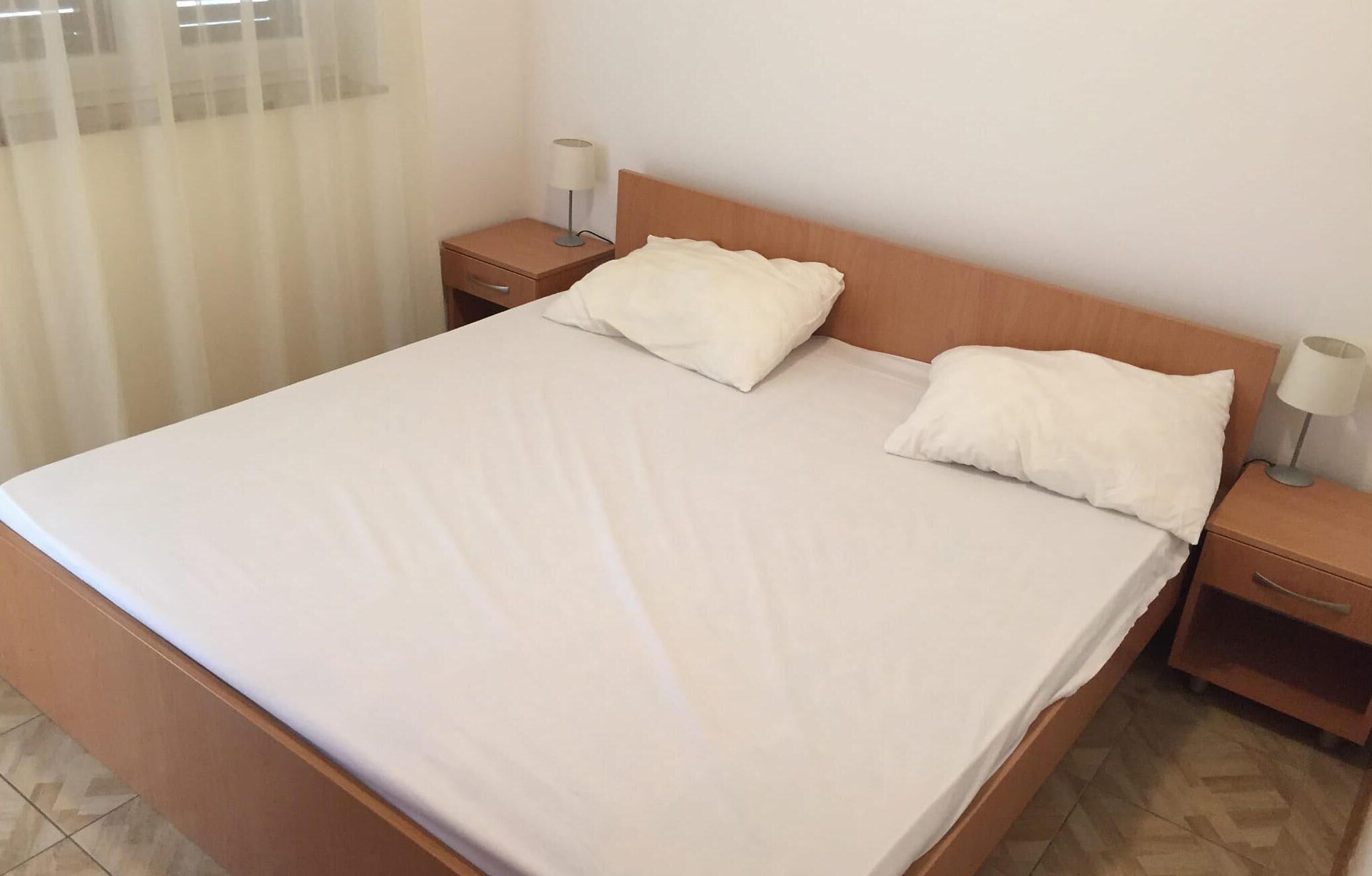 A neatly made bed with white linens and two pillows, wooden headboard, and two bedside tables with lamps.