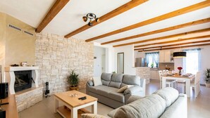 Una sala de estar con chimenea de piedra, vigas de madera, un sofá gris y un comedor con una mesa y sillas blancas.