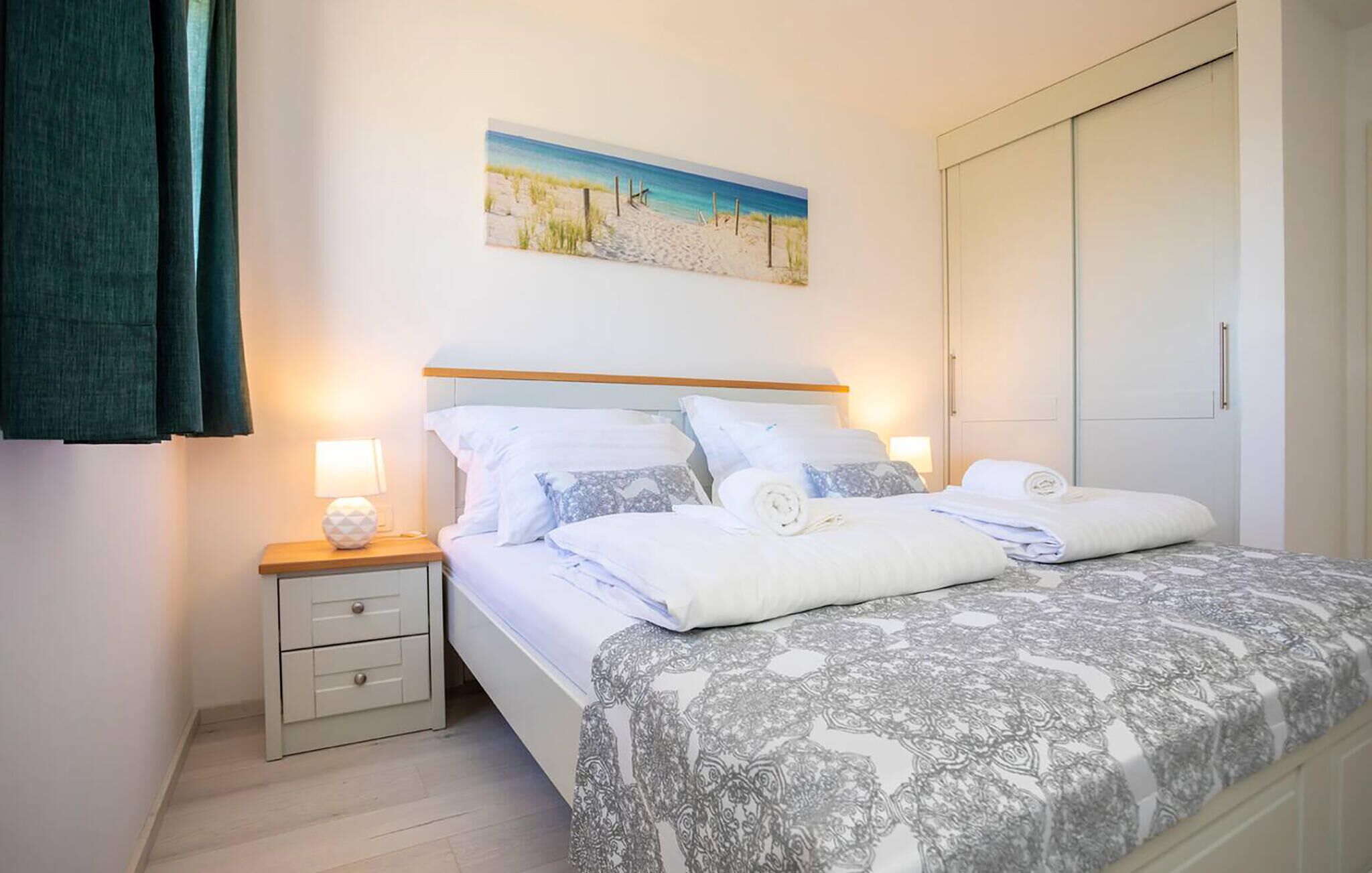 Un lit double bien fait, avec une couvre-lit à motifs, une table de chevet avec une lampe et un tableau représentant une scène de plage accroché au mur.