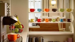 Una cocina con una cesta de fruta, una cafetera y una variedad de tazas coloridas en estantes.