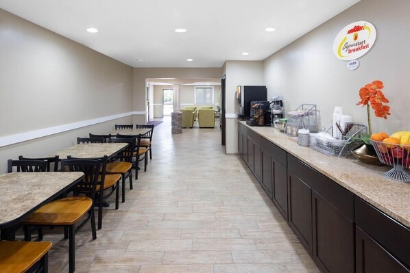 Espaço de refeições de hotel com mesas e cadeiras compridas, balcão de cozinha com opções de café da manhã, e um letreiro com 'Superstart breakfast'.