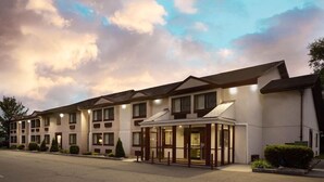 Un hotel de varios pisos con una fila de habitaciones, una entrada central y un estacionamiento.
