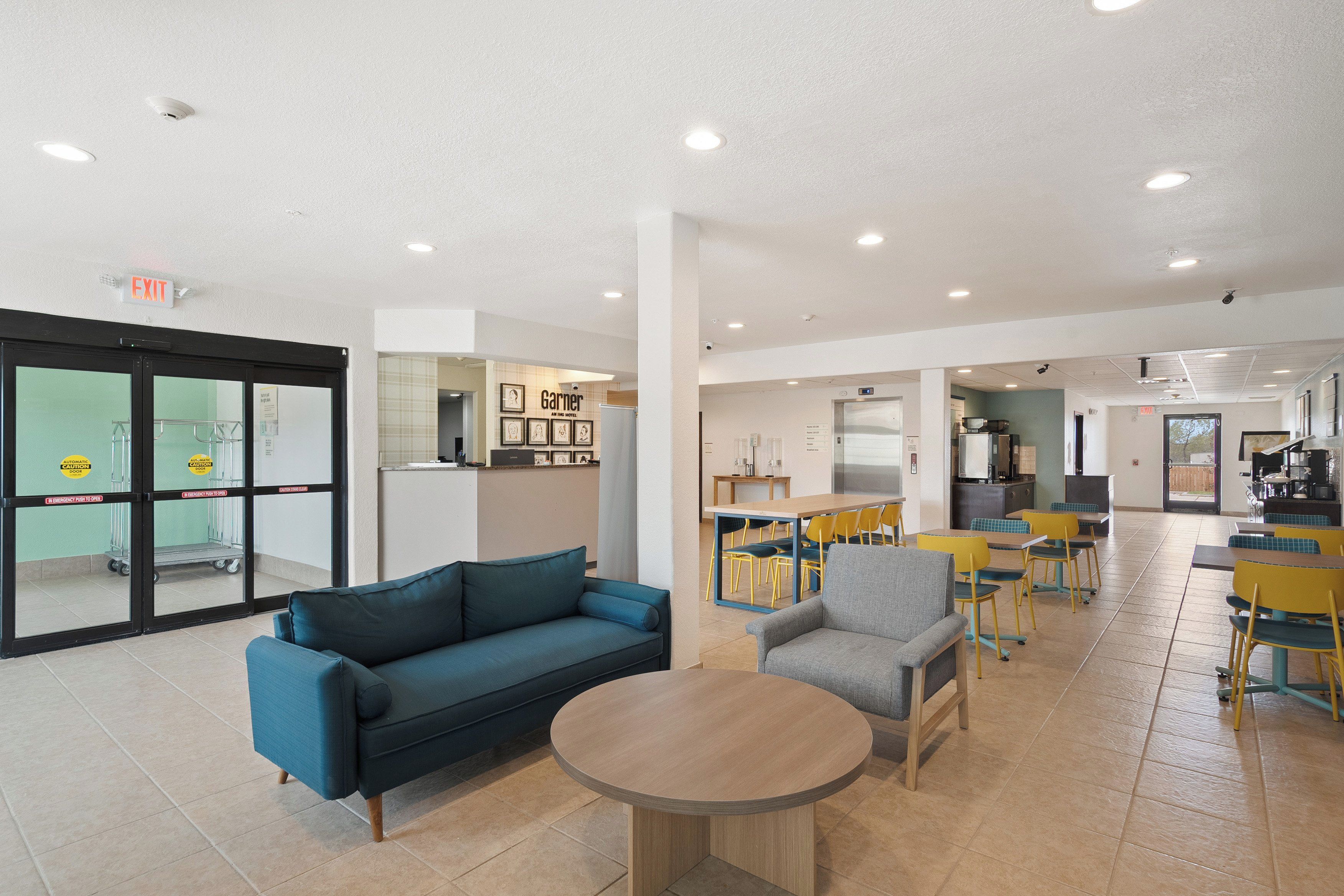 Un vestíbulo amplio con un sofá azul, una mesa redonda y sillas amarillas. Se observa una zona de cocina al fondo con una mesa de comedor y sillas.
