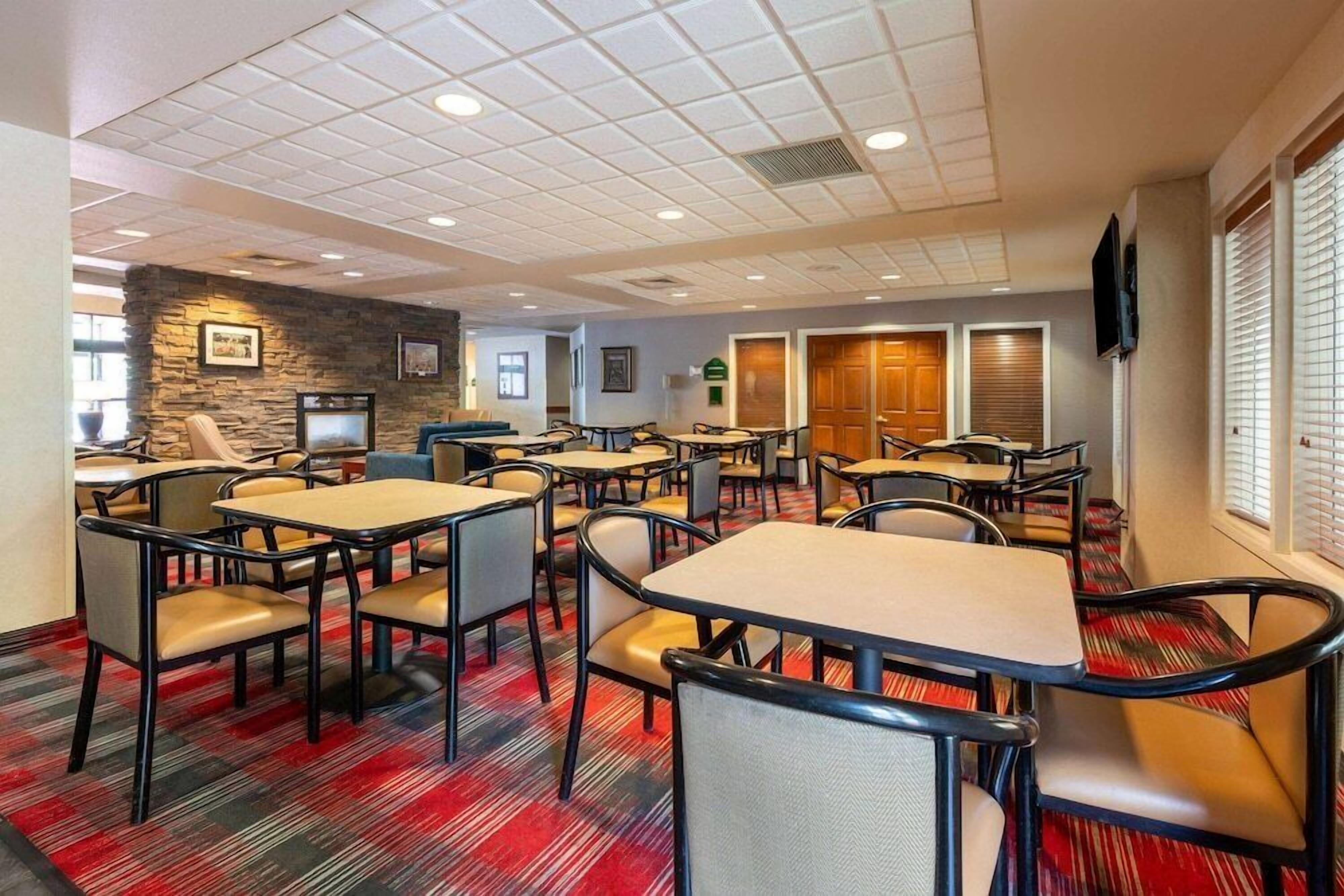 A hotel dining area with multiple tables and chairs, a stone accent wall, and a television mounted on the wall.