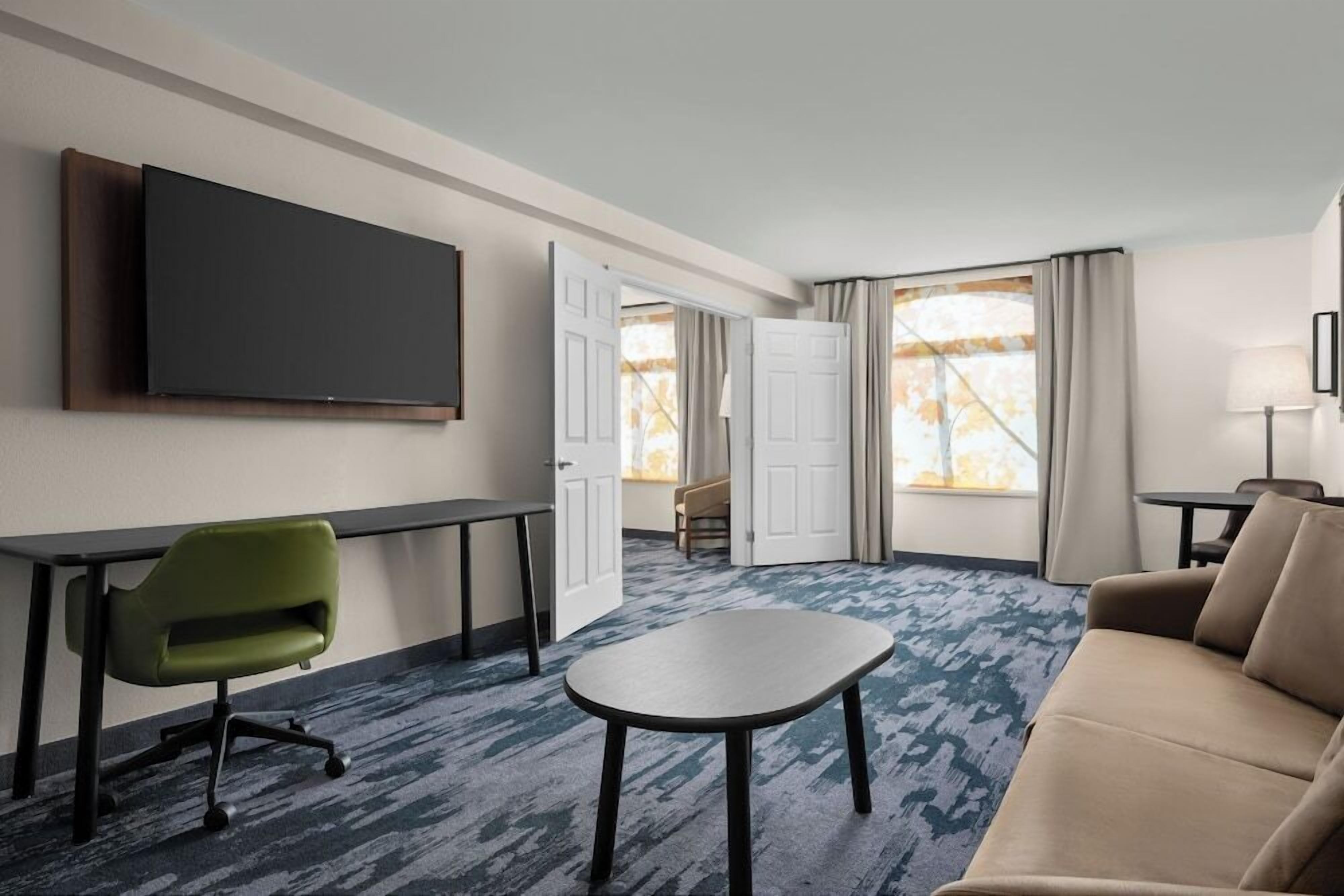 A hotel room with a flat-screen TV mounted on the wall, a green office chair, a small round table, a beige sofa, and a large window with curtains.