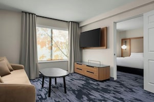 A hotel room with a sofa, a small table, a TV mounted on the wall, a wooden dresser, and a bed with a nightstand.