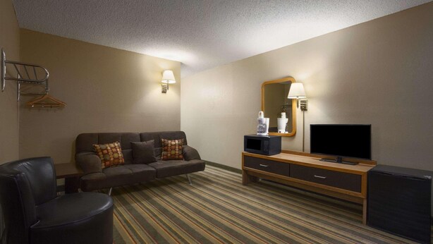 A hotel room with a sofa, a chair, a TV, a mini-fridge, a lamp, and a coat rack. - Super 8 by Wyndham Blackwell (Blackwell)