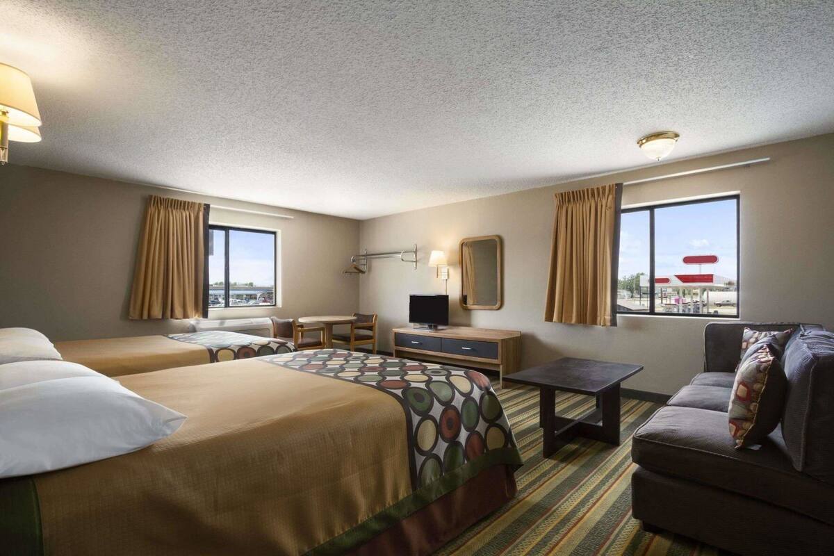 A hotel room with two beds, a desk with a TV, a couch, and large windows with curtains.
