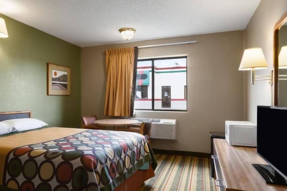 A hotel room with a bed, a desk, a television, a window with curtains, and a wall-mounted lamp.