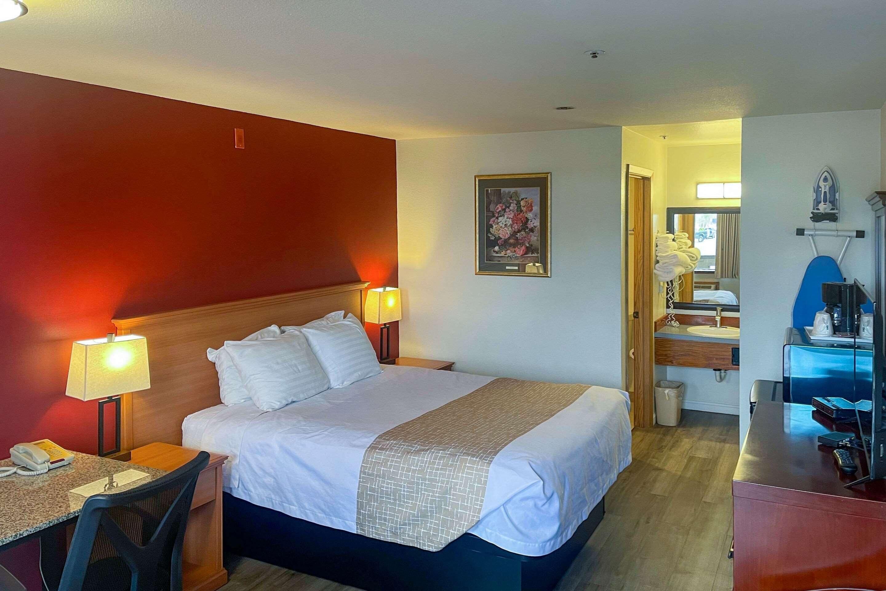 A hotel room with a bed, bedside lamps, a desk with a telephone, a chair, a bathroom with a sink and mirror, and a small desk with a computer.