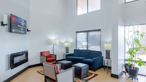 Een moderne woonkamer met een blauwe bank, een rode fauteuil en een tafeltje met lampen. Er is een tv aan de muur bevestigd en een plant in pot bij het raam.