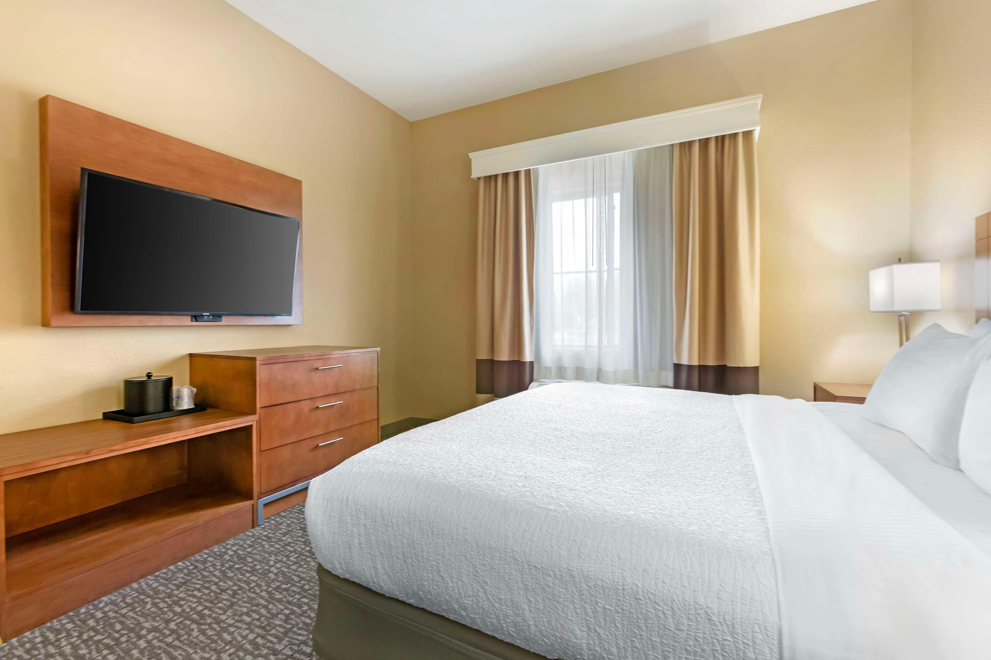 A hotel room with a large bed, a flat-screen TV mounted on the wall, a wooden desk, and a window with curtains.