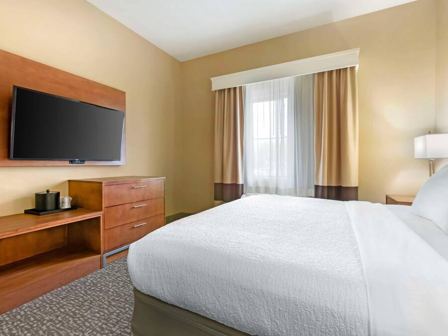 A hotel room with a large bed, a flat-screen TV mounted on the wall, a wooden desk, and a window with curtains.