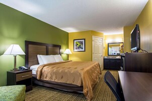 A hotel room with a bed, bedside tables, a desk, a chair, a television, and a kitchen area.