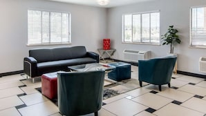 En moderne stue med en sort sofa, to lænestole, et glasbord og en rød dekorativ genstand. Der er to vinduer med persienner, en radiator og en potteplante.