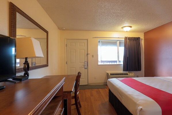 A hotel room with a bed, a desk with a lamp, a television, a window with curtains, and an air conditioning unit. - Travelodge by Wyndham Chehalis (Chehalis)
