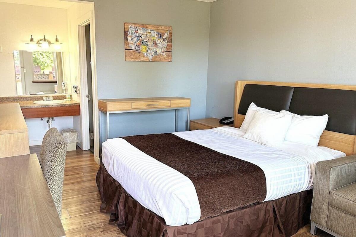 A hotel room with a bed, a desk, a chair, a nightstand, a mirror, and a wall art piece.