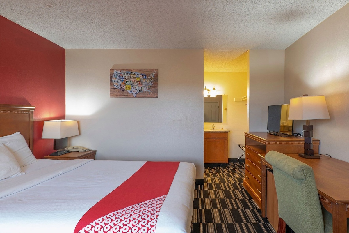 A hotel room with a bed, a desk, a chair, a desk lamp, a telephone, a painting on the wall, and a bathroom visible through an open door.