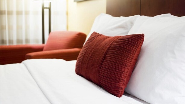 Un lit bien fait, avec des draps blancs et un seul oreiller rouge.