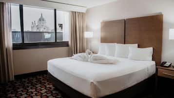 A hotel room with a large bed, a nightstand, a lamp, and a view of a building through the window.