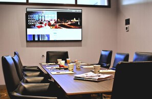 A conference room with a long table, chairs, a screen displaying an event center, and a sign indicating maximum occupancy.