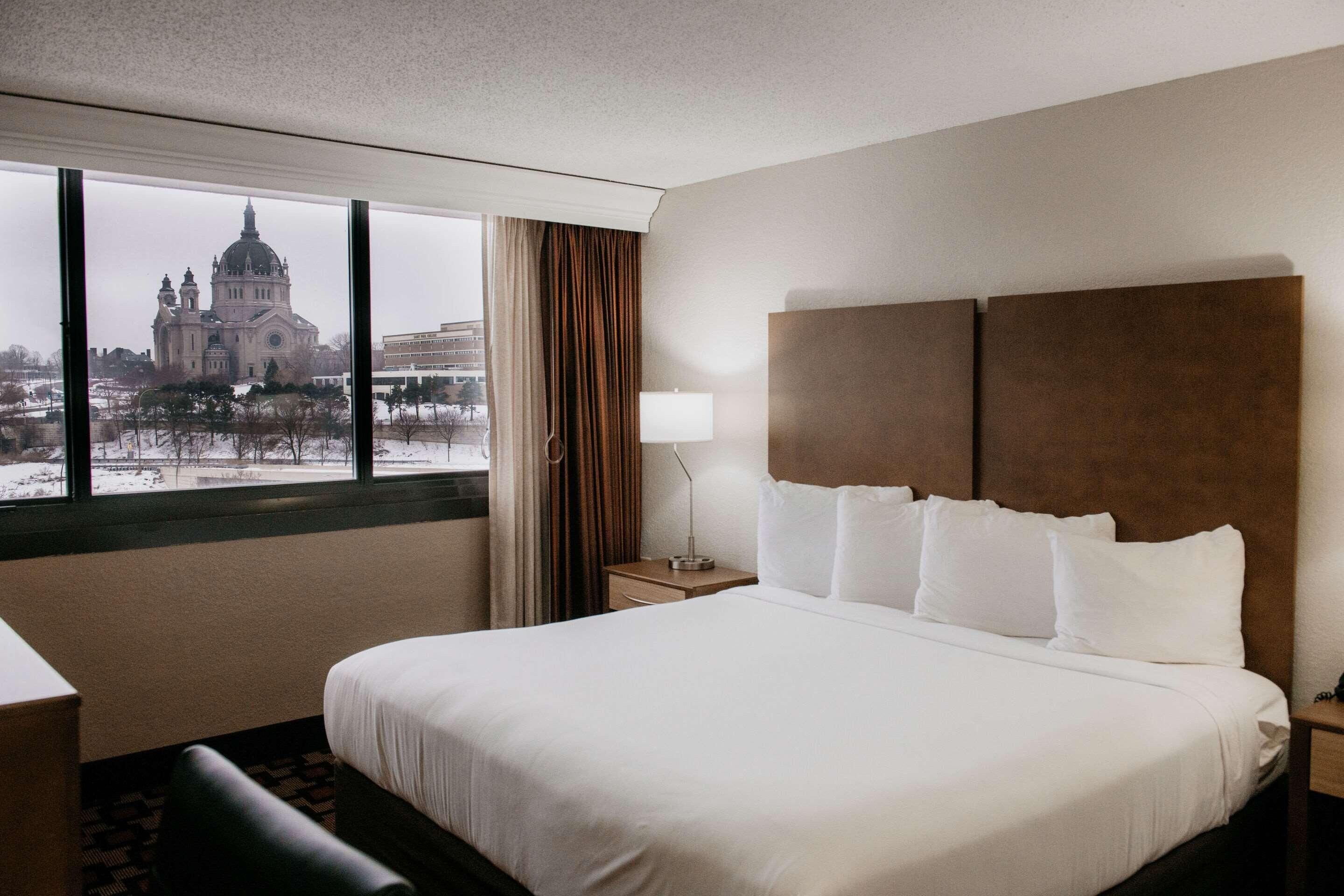 Hotellihuone, jossa on suuri sänky, yöpöytä, lamppu ja ikkunasta avautuva näkymä katedraaliin.