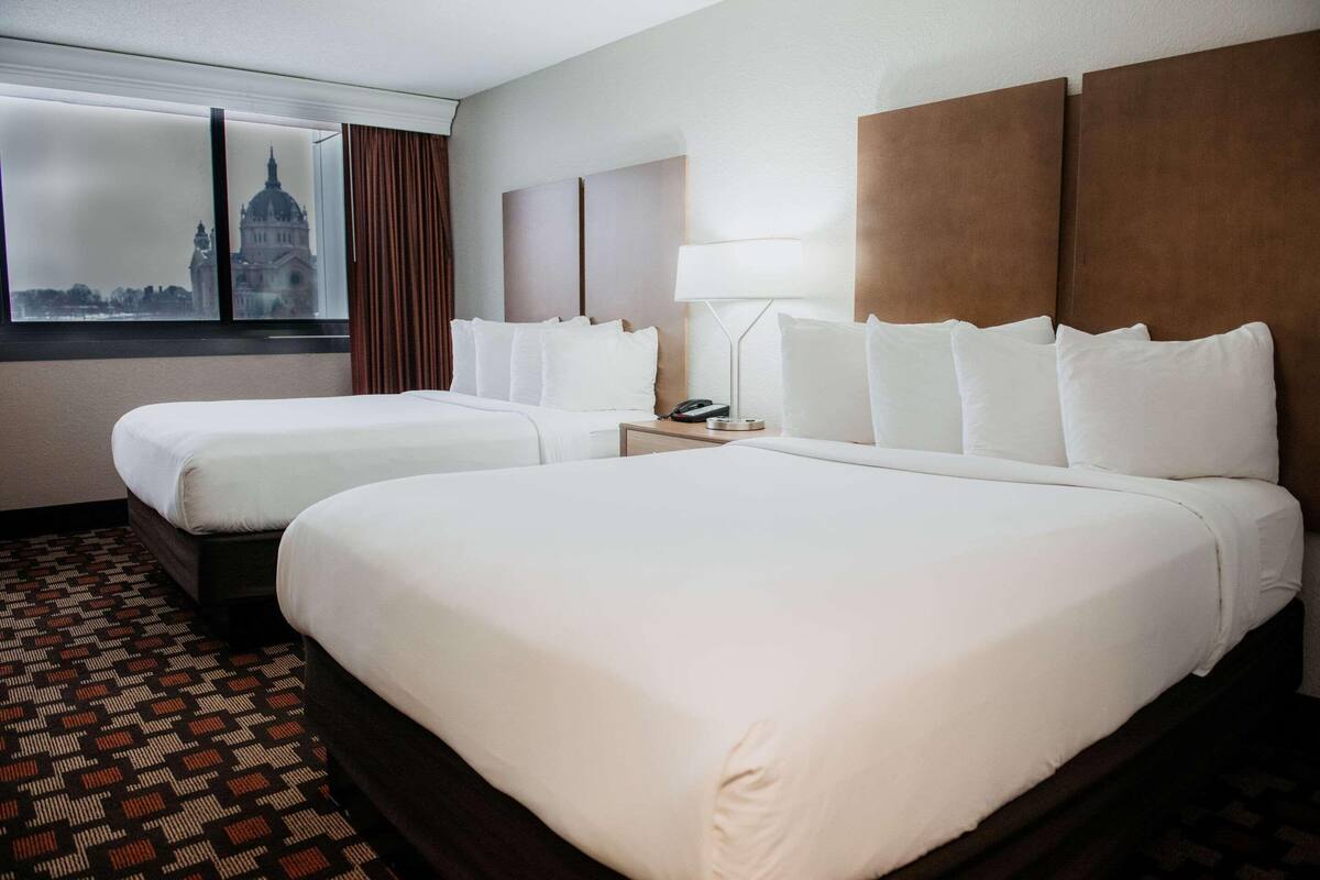 A hotel room with two beds, a nightstand with a lamp, and a window with a view of a building.